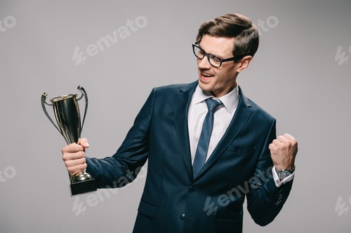 Preview: cheerful businessman in glasses celebrating victory and holding trophy in hand on grey background