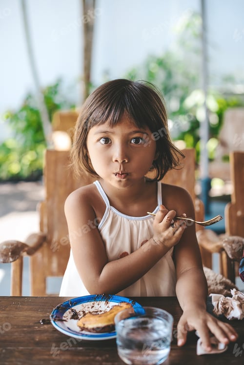 Preview: Funny little girl looks at camera eating dessert at table in cafe
