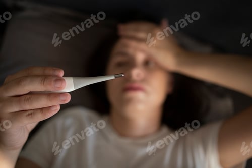 Preview: woman lying on the bed having high fever and measuring her temperature with a thermometer