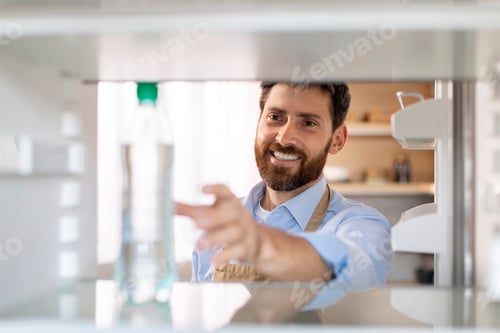 Preview: Smiling millennial caucasian bearded guy in apron opens door of empty refrigerator and takes bottle