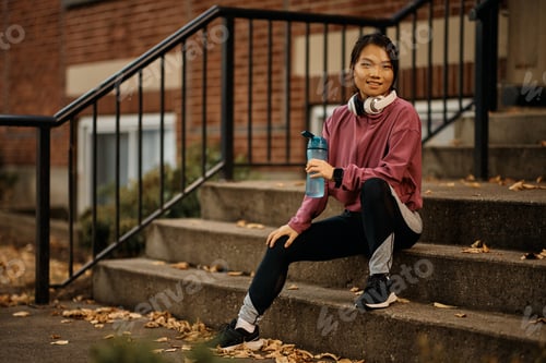 Preview: Happy Asian athlete relaxing during her water break outdoors and looking at camera.