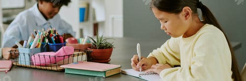 Preview: Asian Girl Writing in Notebook While Black Boy Drawing in Classroom Setting