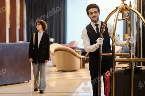 Preview: Young elegant bellboy in suit pushing cart with many suitcases