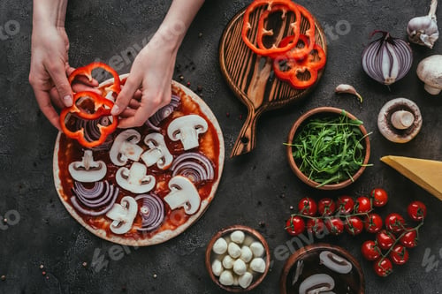 Preview: cropped shot of woman spreading bell pepper slices onto pizza on concrete table