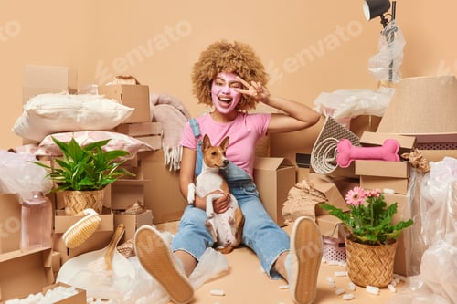 Preview: Happy young woman with curly hair applies beauty facial mask makes peace gesture winks eye spends ti
