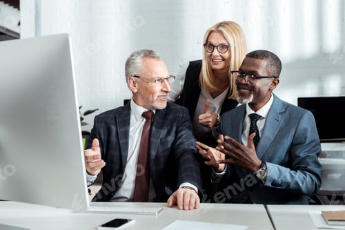 Preview: happy blonde woman in glasses showing thumb up near multicultural partners