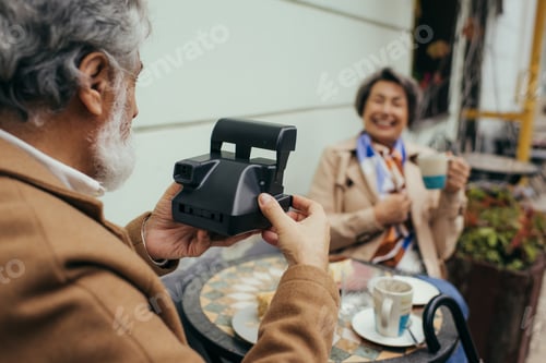 Preview: bearded man holding vintage camera and taking photo of blurred wife during breakfast