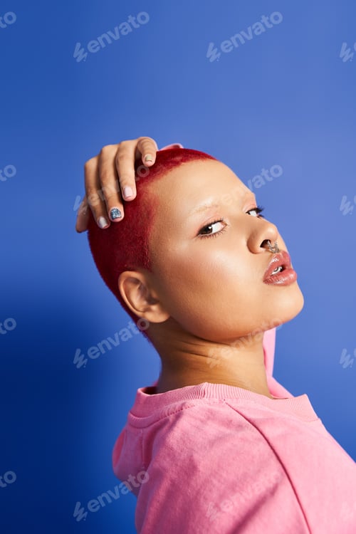 Preview: A young woman with short pink hair and a nose piercing poses in a vibrant studio