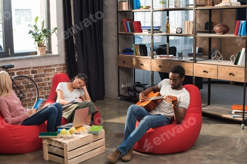 Preview: african american male student playing guitar while girls studying with books near