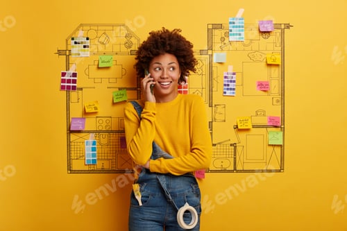 Preview: Happy ethnic woman designer speaks via cell phone, looks away, stands against house planning on yell