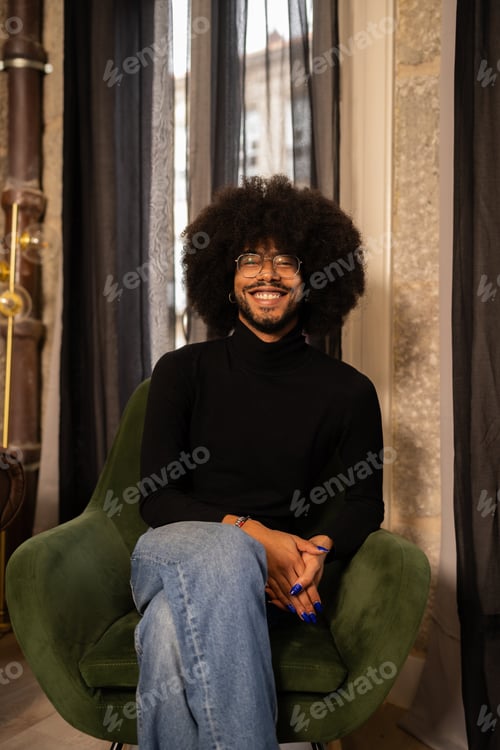 Preview: Non binary person with afro hair, painted nails, smiling while sitting on green armchair