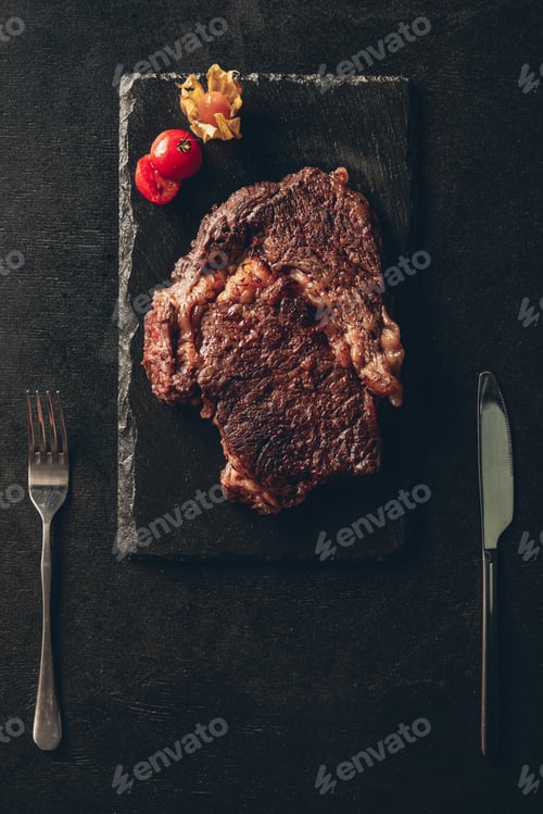 Preview: top view of cooked steak and cherry tomato on black wooden board, knife and fork on table in kitchen