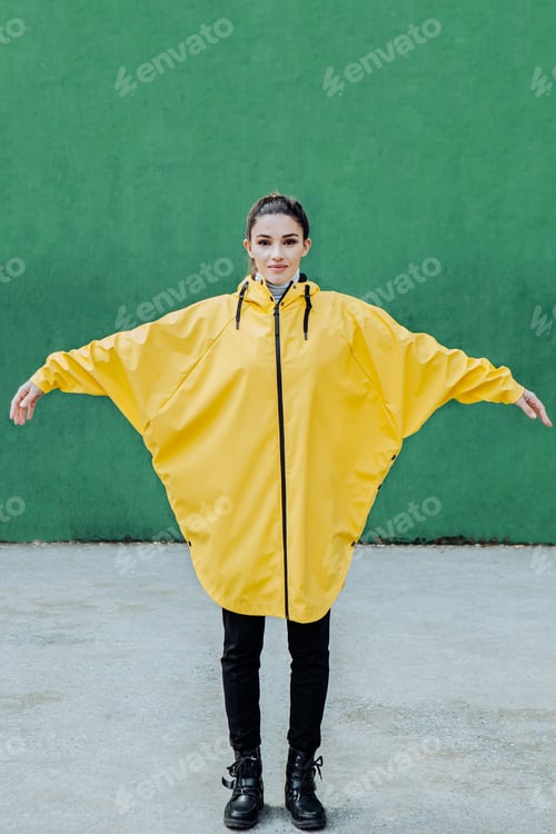 Preview: A millennial girl dressed in a yellow raincoat poses in front of a green wall.