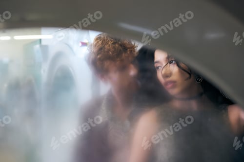 Preview: interracial couple in stylish outfits standing behind glass of washing machine in coin laundry