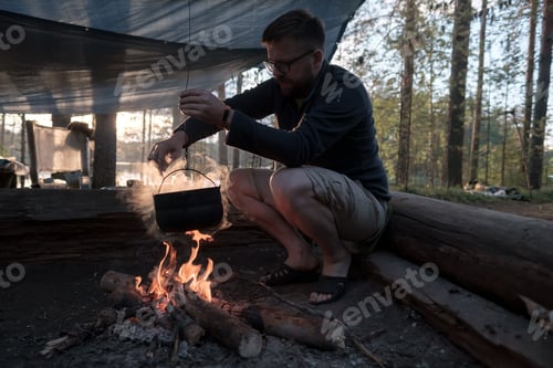 Preview: Tourist cooking dinner over campfire at camp, stirring food in bowler over fire