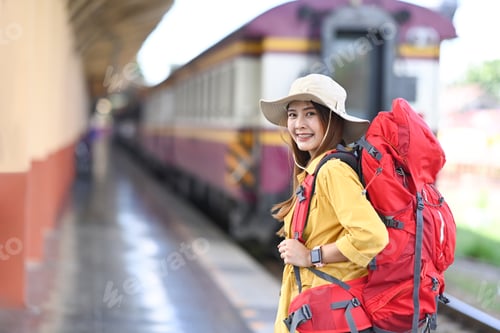 Preview: Cheerful young asian woman with backpack smiling to camera while standing in train station.