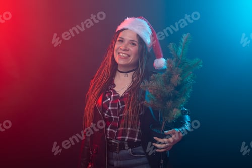 Preview: Christmas, rock, studio, people concept - young woman in christmas hat with small christmas tree