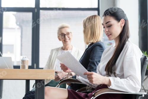 Preview: side view of beautiful businesswomen reading documents and talking at meeting in office