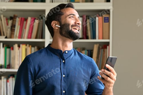 Preview: Happy indian business man at work wearing earbud using smartphone.