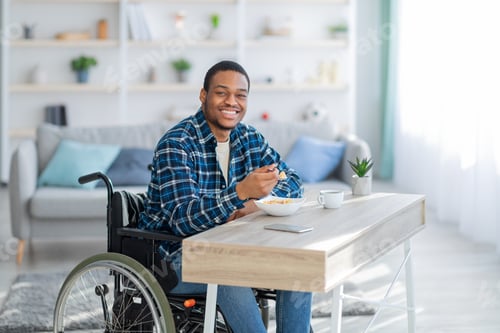 Preview: Cheerful impaired black guy in wheelchair having breakfast, eating tasty cereal at home