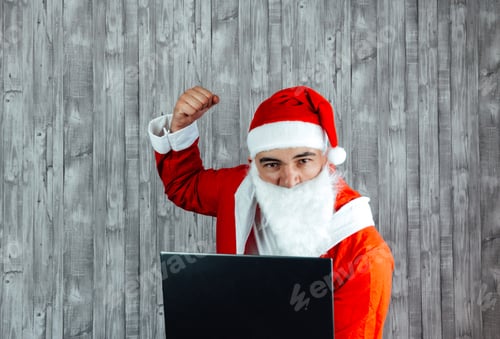 Preview: Young Caucasian man dressed as Santa Claus angry with fist up with computer in front of him