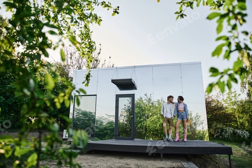 Preview: full length of happy and stylish interracial couple standing on porch of glass house in countryside