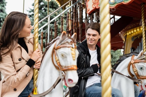 Preview: cheerful young man looking at girlfriend riding carousel horse in amusement park