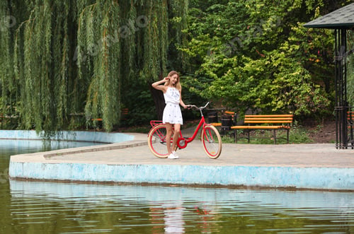 Preview: Beautiful woman in casual outfit with bicycle near pond outdoors