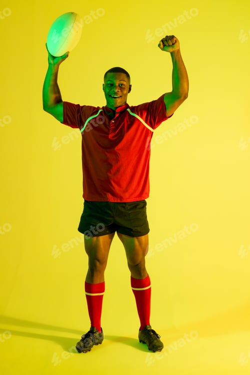 Preview: Portrait of happy african american male rugby player raising hands over yellow lighting