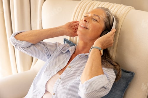 Preview: Relaxing at home, mature woman enjoying music with headphones on chair