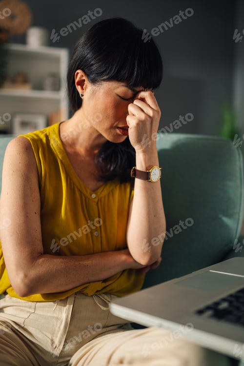 Preview: Asian woman in a yellow top sitting on couch with laptop, thinking.