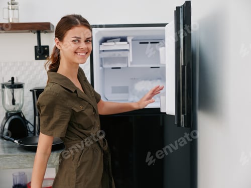 Preview: Woman smiling with teeth looking into camera in kitchen at home opened freezer empty with ice inside