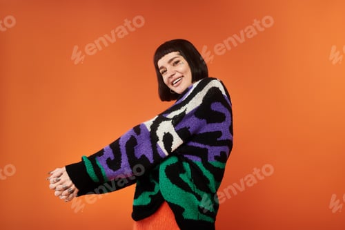 Preview: Young woman with tattoos poses in vibrant sweater against bright orange studio backdrop