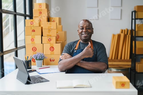 Preview: Smiling Small Business Owner in Apron Giving Thumbs Up in Modern Office with Cardboard Boxes and Lap