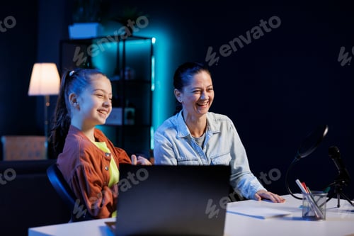 Preview: Daughter and mother filming reaction video for gen Z audience, looking at online videos on laptop
