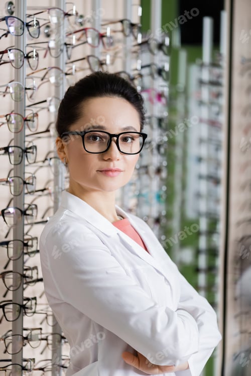 Preview: oculist in white coat standing with crossed arms near assortment of eyeglasses in optics shop