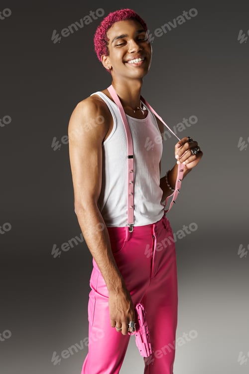 Preview: joyful young man in stylish attire posing with pink toy gun and smiling cheerfully at camera