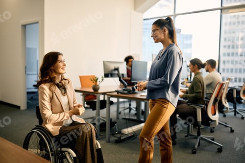 Preview: Happy businesswoman in wheelchair talking to female colleague in the office.