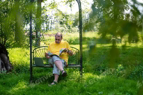Preview: middle aged man with laptop and documents working outside in garden, green home office concept.