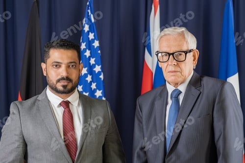 Preview: Two elegant male delegates from various countries standing against flags and looking at camera
