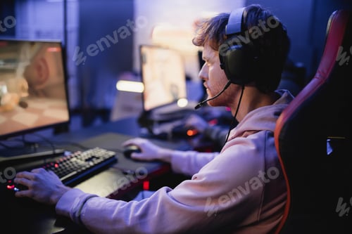 Preview: Side view of young gamer in headphones playing computer game in cyber club
