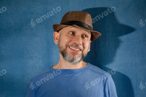 Preview: Caucasian male wearing a blue t-shirt and a hat standing against a blue wall smiling happily