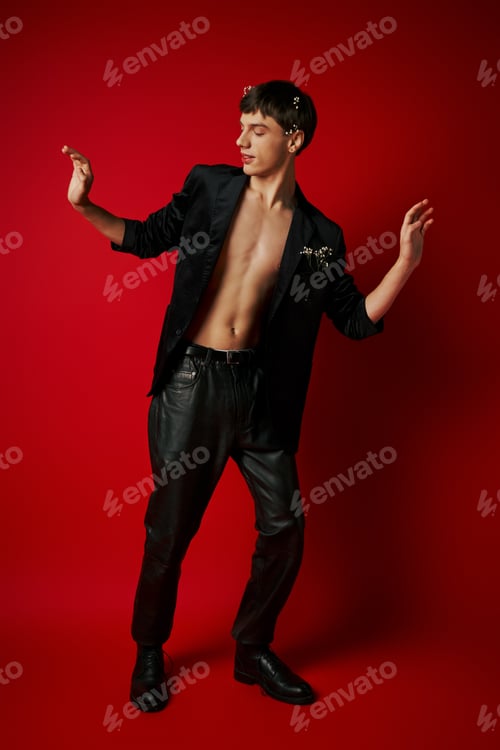 Preview: full length of trendy young man in black attire with flowers in his hair gesturing on red background