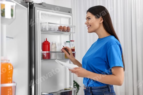Preview: Young woman with notebook and pen near modern refrigerator indoors
