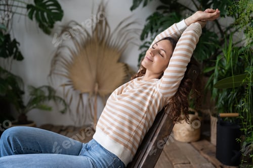 Preview: Serene relaxed young woman with closed eyes stretching arms, relaxing in urban jungle interior