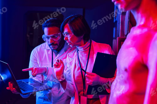 Preview: indian scientist pointing at laptop near colleague and humanoid alien in science center