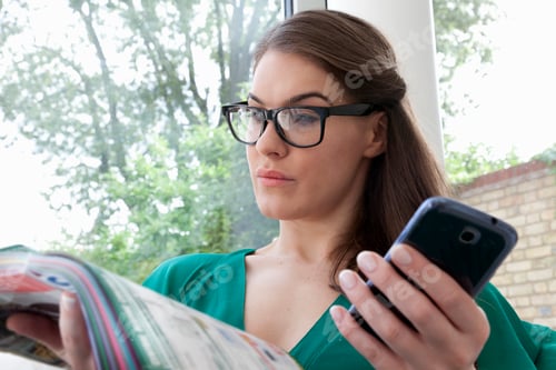 Preview: Young woman wearing glasses reading magazine holding cell phone