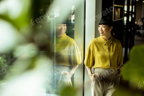 Preview: Woman in yellow blouse stands by large window in a modern workspace during daylight