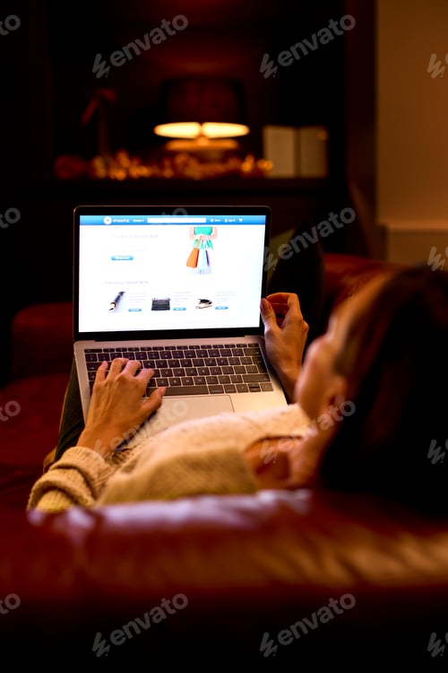 Preview: Rear View Of Woman At Home Sitting In Lounge At Night Shopping Online