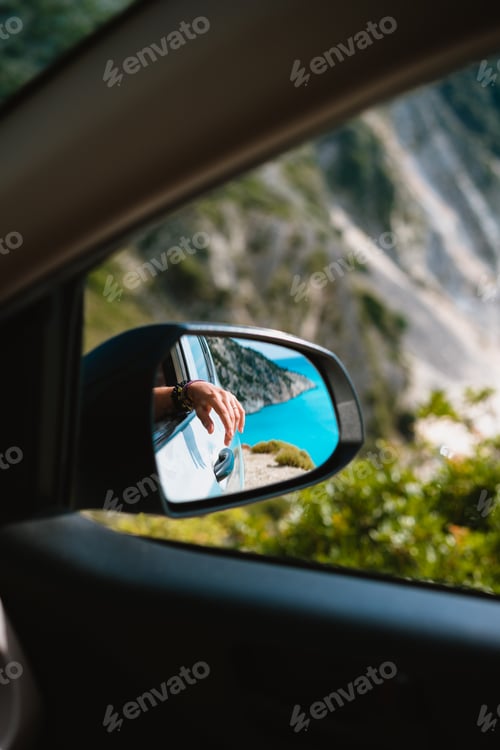 Preview: Female hand mirrored in the car side view mirror. Blue mediterranean sea and white rocks in